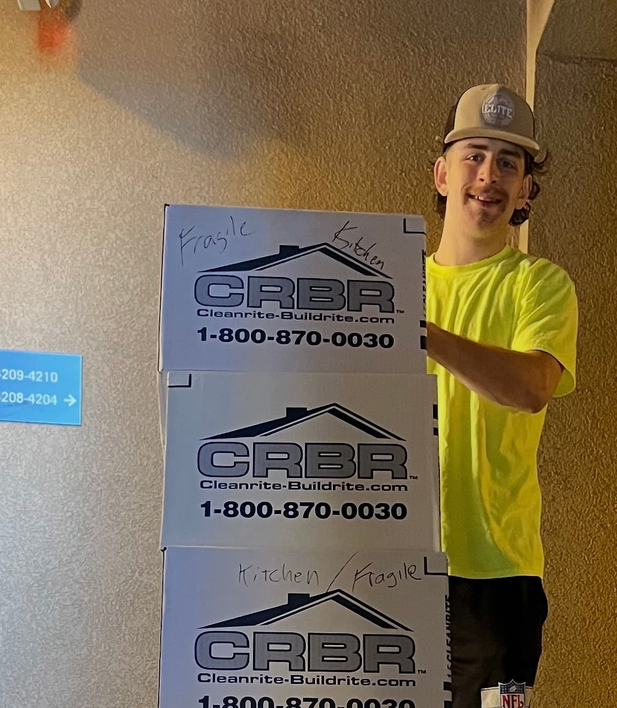 Smiling man in yellow shirt stands behind three stacked CRBR boxes labeled kitchen and fragile.