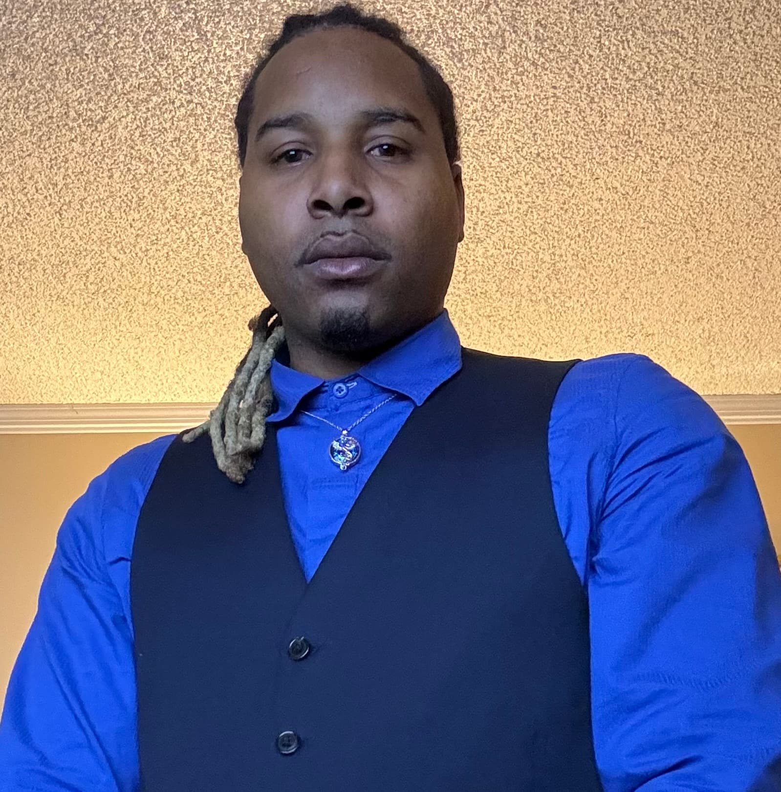 Black man with locs wearing a royal blue shirt and black vest with a pendant.