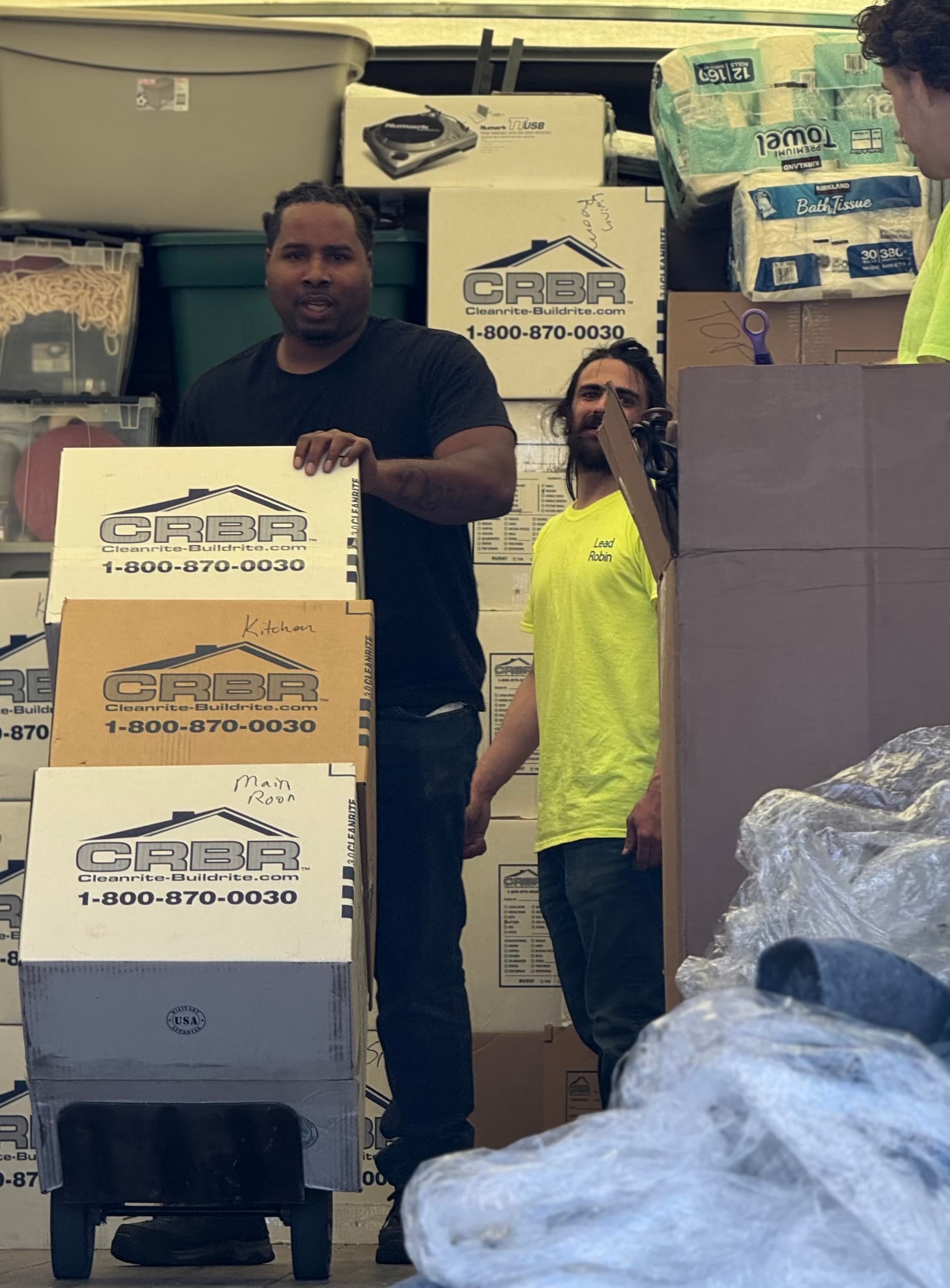 Two movers stand in a packed truck with boxes labeled CRBR on a dolly.